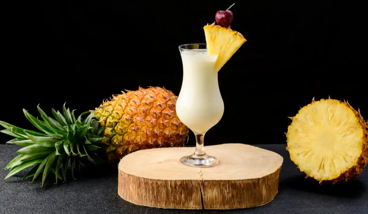 A glass with pina colada cocktail, a whole pineapple next to the drinks on a black background.