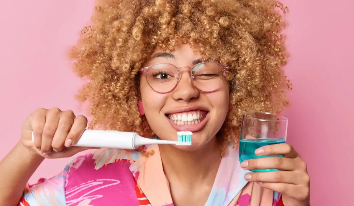 Happy young woman holds glass of fresh mouthwash and electric toothbrush winks